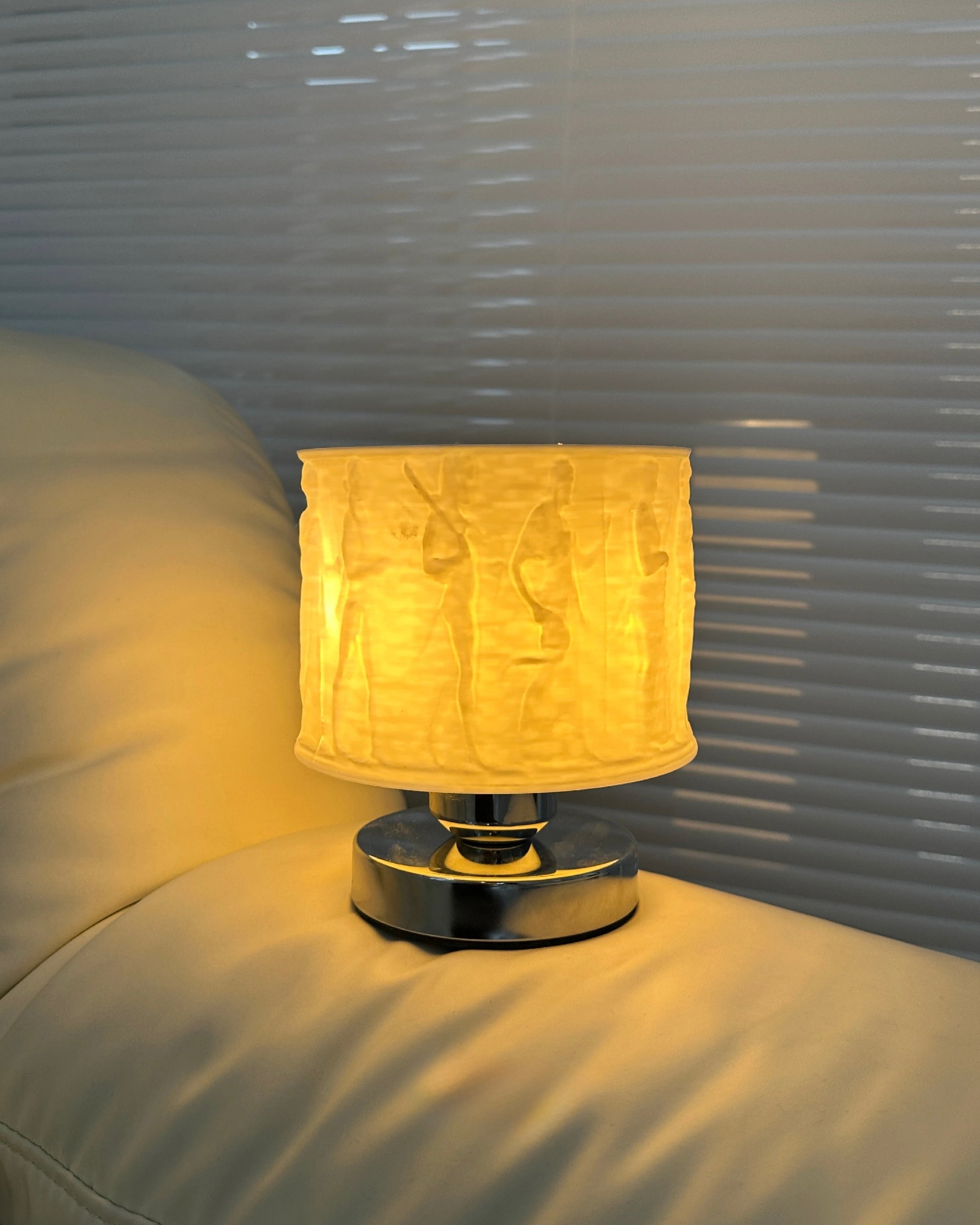 Small table lamp with a textured shade on a bed next to a window with blinds.