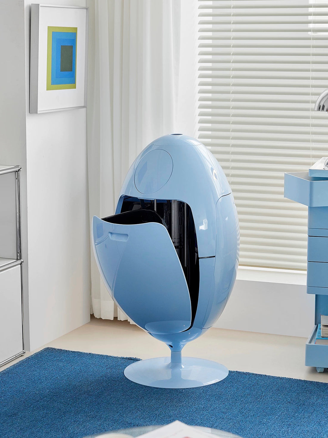 Blue egg-shaped chair in a room with a window and artwork on the wall.