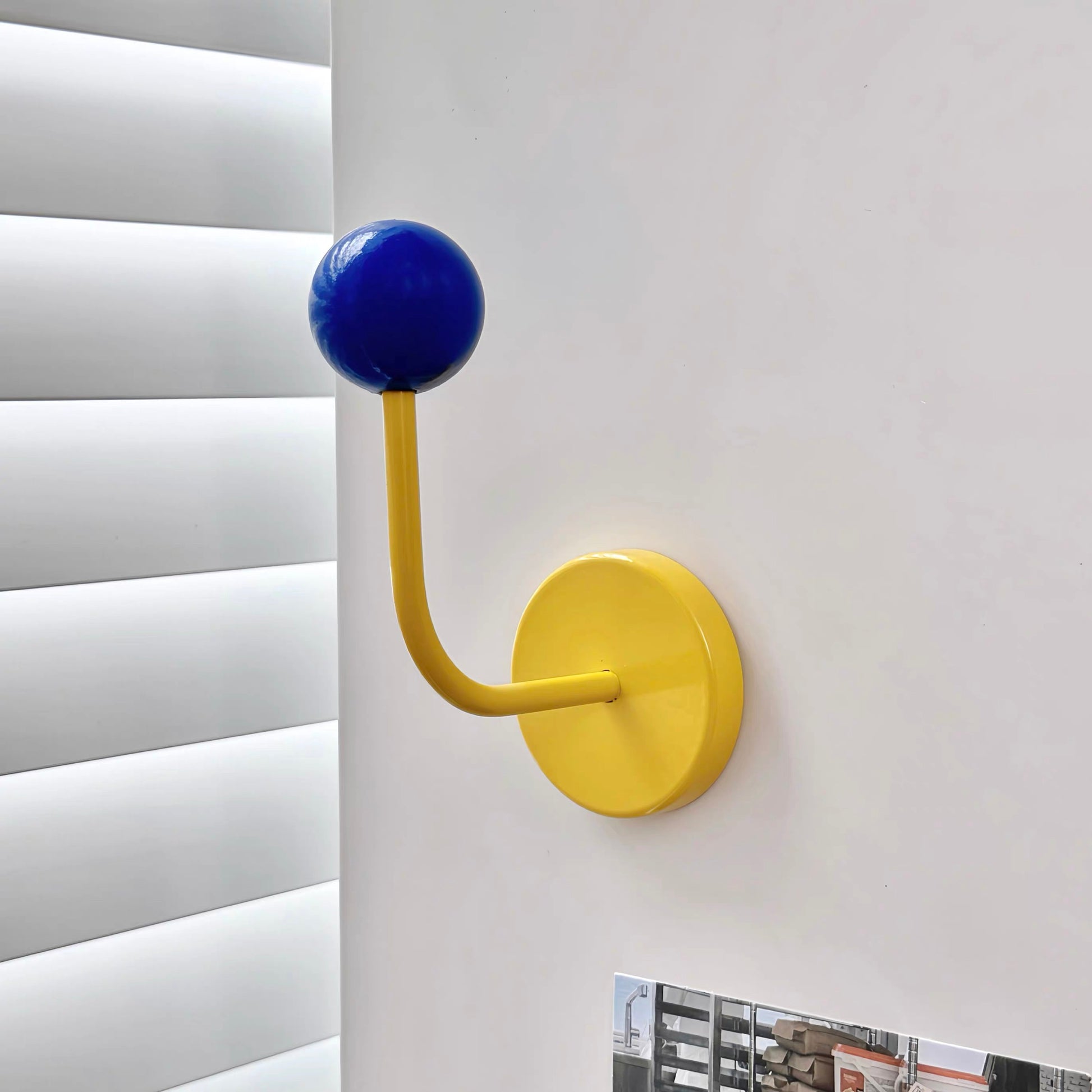A modern wall-mounted coat rack hook with a bright yellow curved stem and circular base, topped with a glossy blue spherical hook, shown mounted on a white wall next to window blinds.