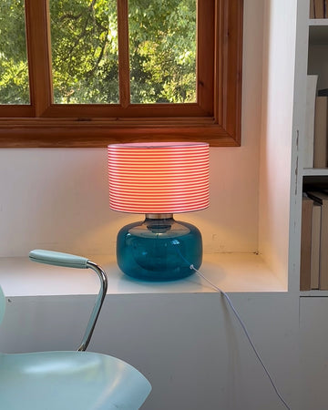 Table lamp with a blue base and pink striped shade on a white surface near a window.
