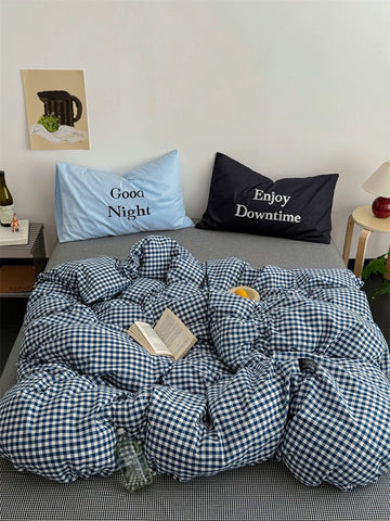 Bed with checkered duvet, pillows labeled 'Good Night' and 'Enjoy Downtime', and a small table with a bottle and vase.