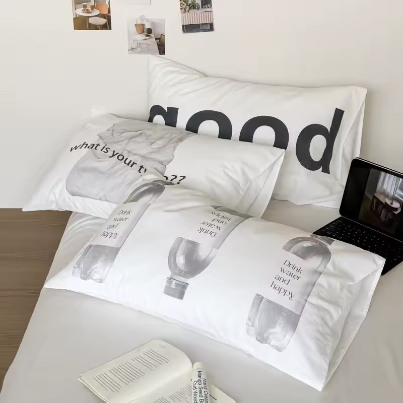 A set of white bedding with black text and water bottle illustrations, styled on a bed with a laptop, open book, and wall photos in the background.