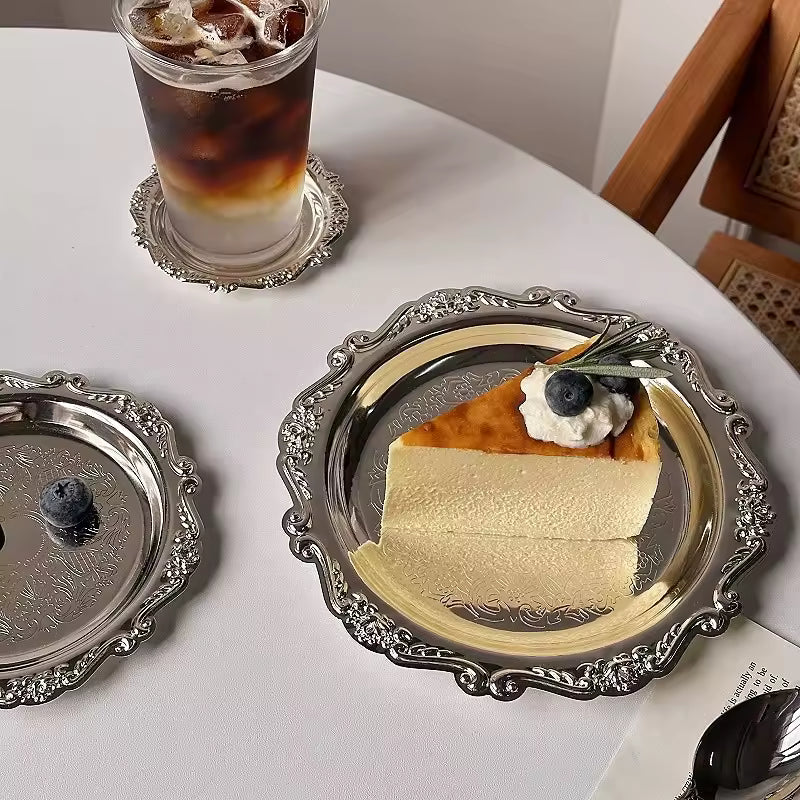 Slice of cheesecake on a decorative silver plate with a glass of iced coffee on a white table.