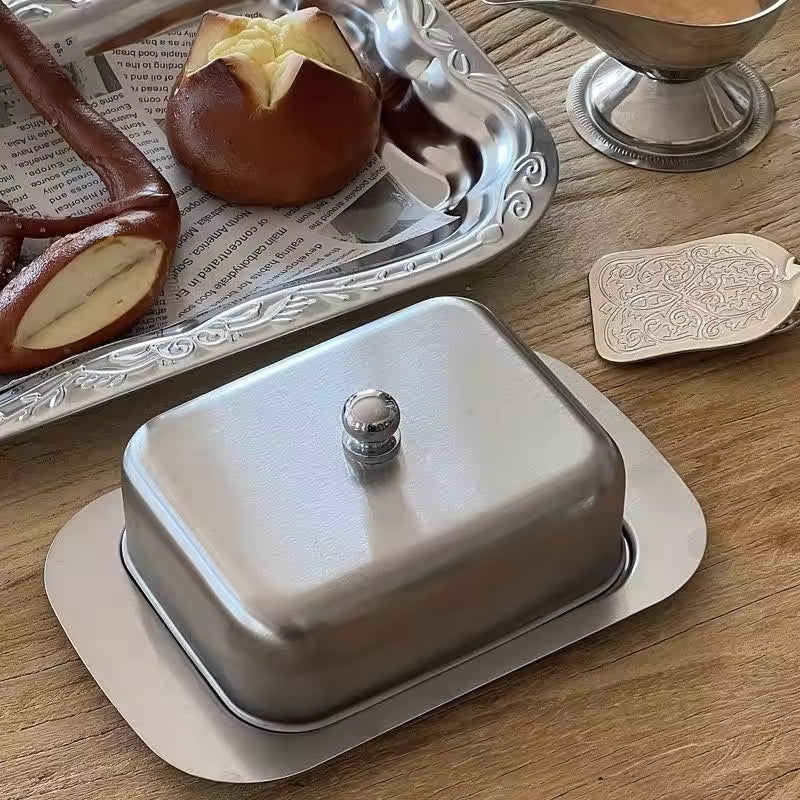 Metallic butter dish with lid on a wooden surface, next to a tray with bread and a small container.