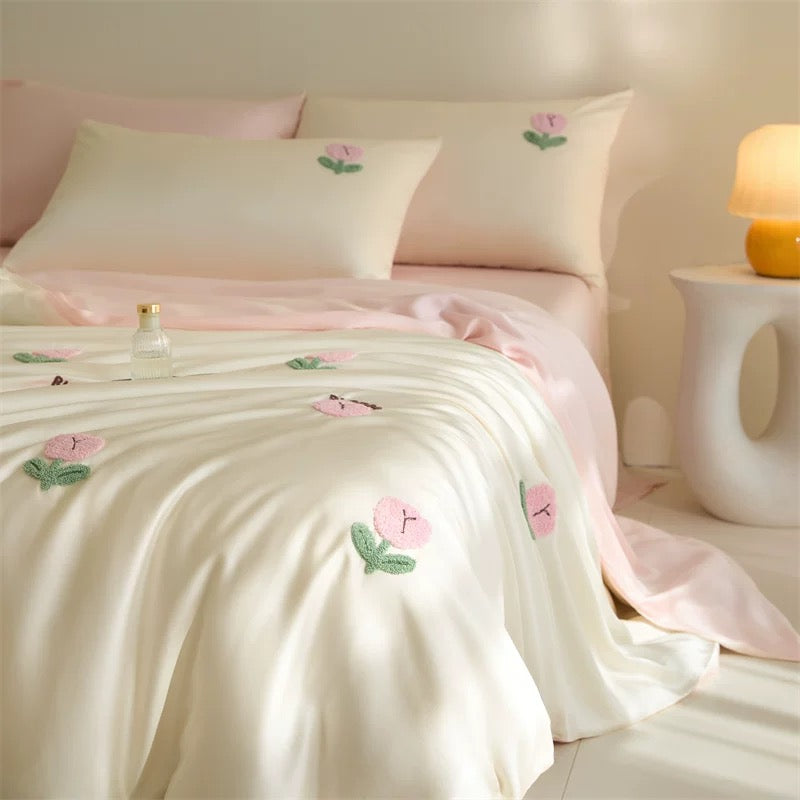 A neatly made bed with a soft cream bedding set and pale pink sheets, decorated with embroidered pink flowers and green leaves. Several matching pillows are arranged on the bed, with a small decorative tray and a lit lamp visible in a softly lit bedroom setting.