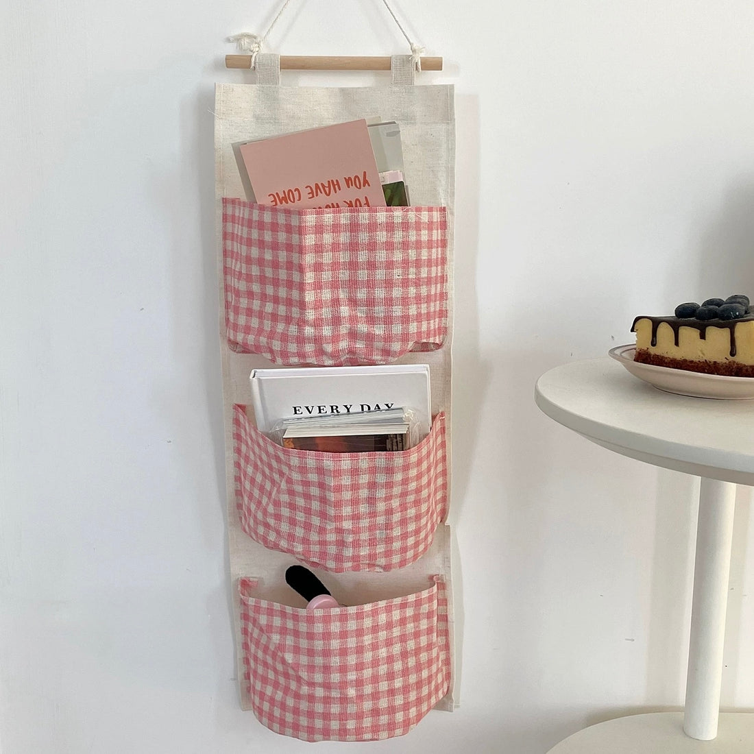 Hanging organizer with pink gingham design on a white wall next to a table with a cake.