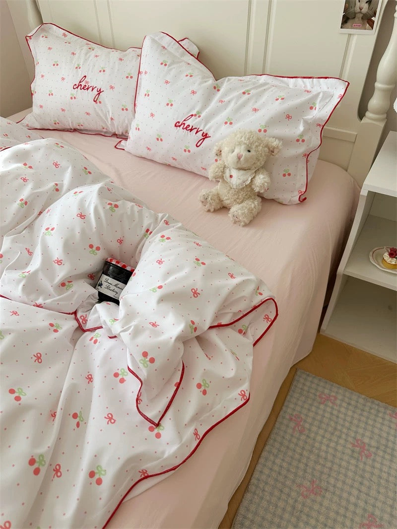 Bed with cherry-themed bedding and a teddy bear, set against a wooden floor.