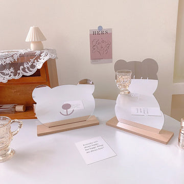 Two acrylic bear-shaped tabletop decor pieces with wooden bases on a white table, styled in a soft indoor setting with a small lamp, lace cloth, and glassware.