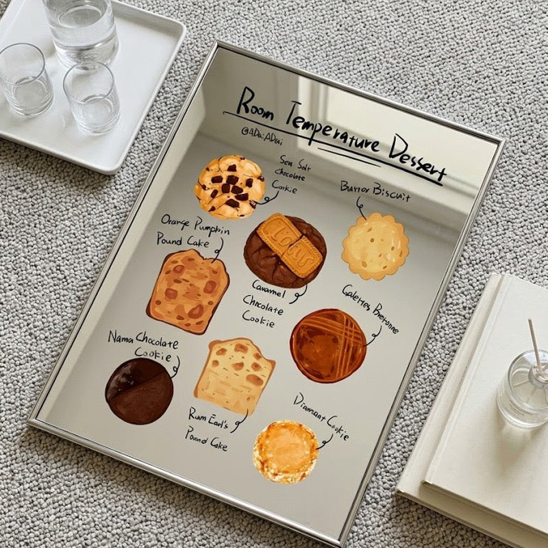 A rectangular framed print featuring a dessert-themed illustration titled 'Room Temperature Dessert' with labeled cookies, cakes, and pastries, displayed on a textured gray surface with books and glassware nearby.