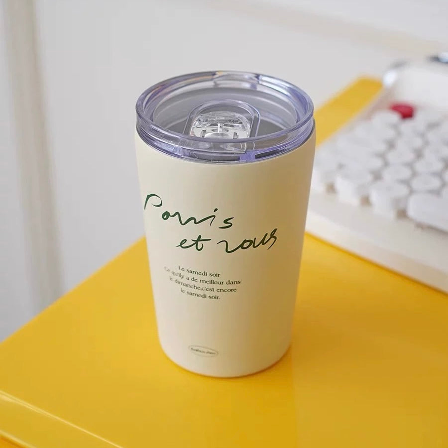 A cream-colored insulated tumbler with a clear lid sits on a bright yellow desk next to a white keyboard. The tumbler features the French phrase 'Romant et nous' printed in green and black script.