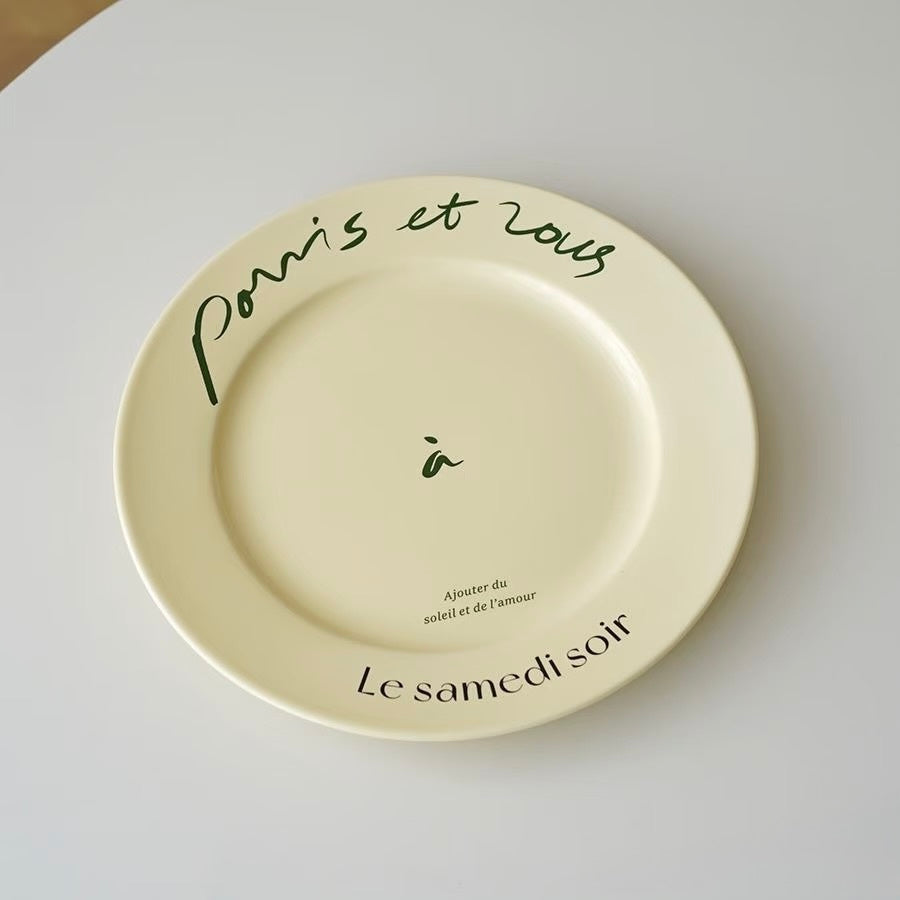 A round cream-colored ceramic plate with French text printed on it, photographed from above on a light gray surface.