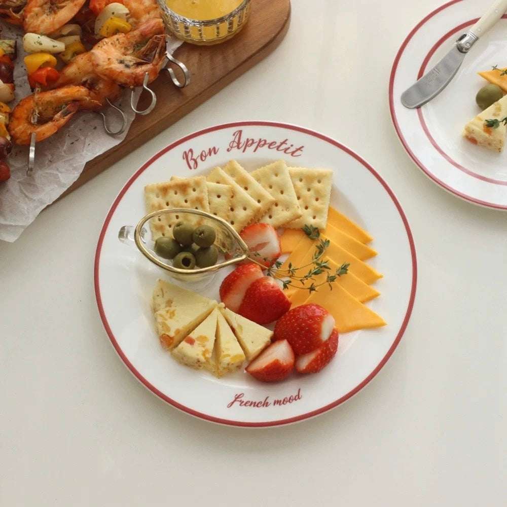 A white ceramic serving platter with red borders and the words 'Bon Appetit' and 'French mood' printed on it, styled with crackers, cheese, strawberries, olives, and a small dipping dish on a white tabletop. A second plate and a wooden serving board with shrimp are partially visible in the background.