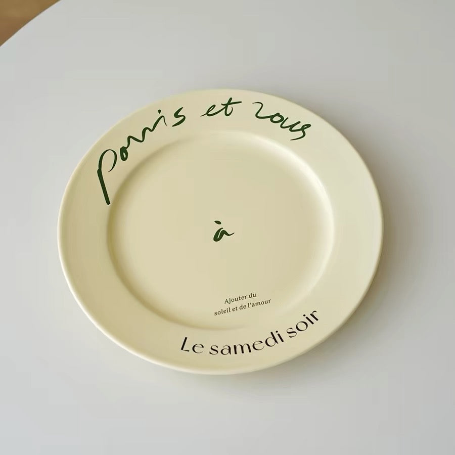 A round cream-colored ceramic plate with French text, photographed on a neutral light gray surface. The plate features 'Paris et Roue' in dark green script on the top rim, a small green 'a' in the center, and 'Le samedi soir' in black on the bottom rim.