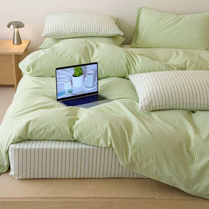 A bed dressed in light green bedding with matching pillows and a striped pillow, set in a minimalist room with a wooden bed frame, side table, and laptop on the mattress.