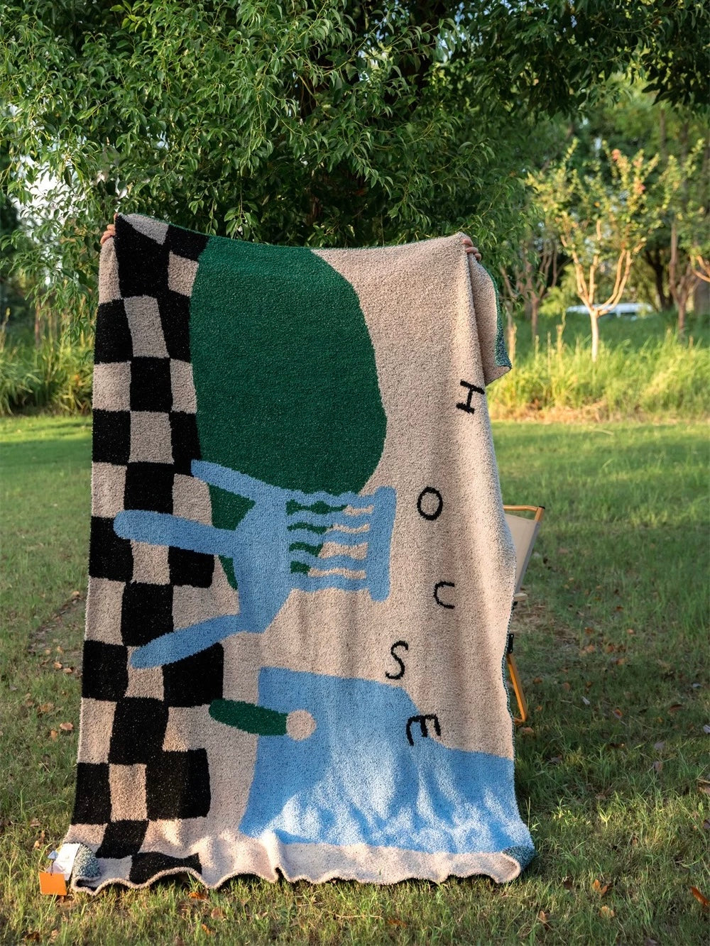 A large woven throw blanket with an abstract geometric pattern in beige, black, green, blue, and white, draped over a chair in an outdoor grassy garden setting.