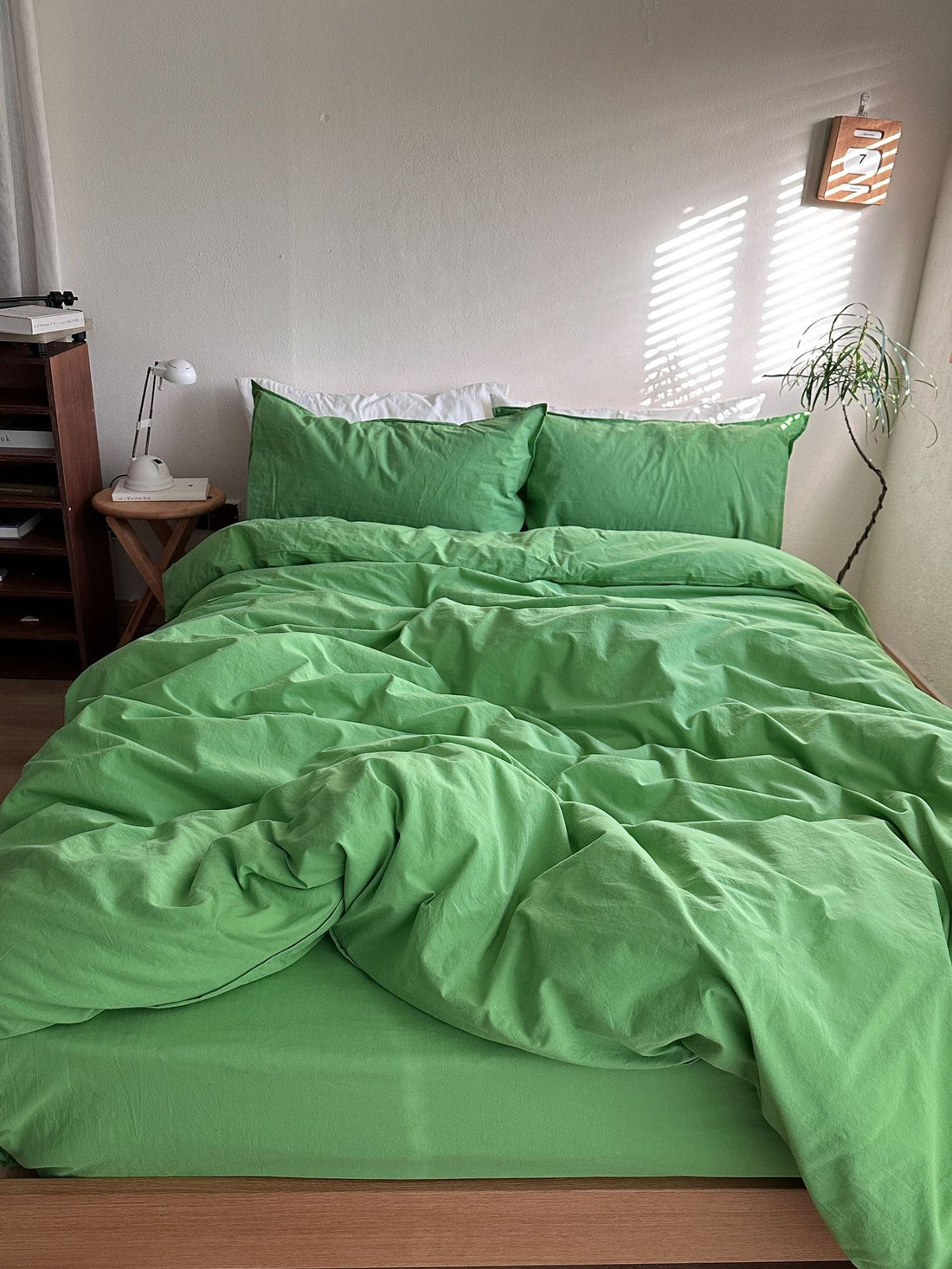 A bed dressed in a bright green bedding set with matching pillowcases and duvet, styled in a minimalist bedroom with a wooden side table, white lamp, and soft window light casting shadows.