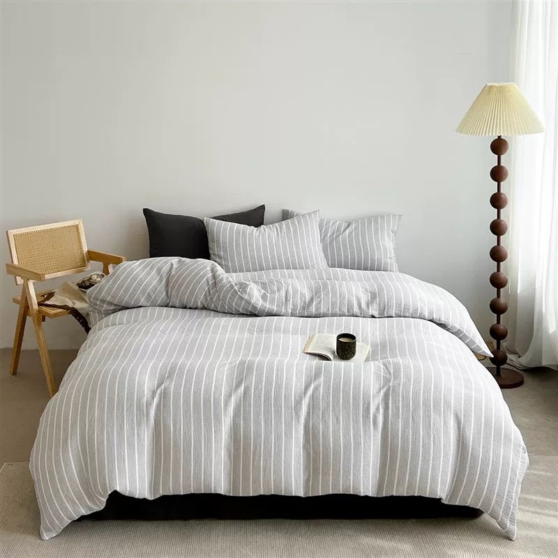 A bed dressed in a light gray and white vertically striped bedding set, styled in a minimalist bedroom with a wooden chair, floor lamp, and neutral wall.