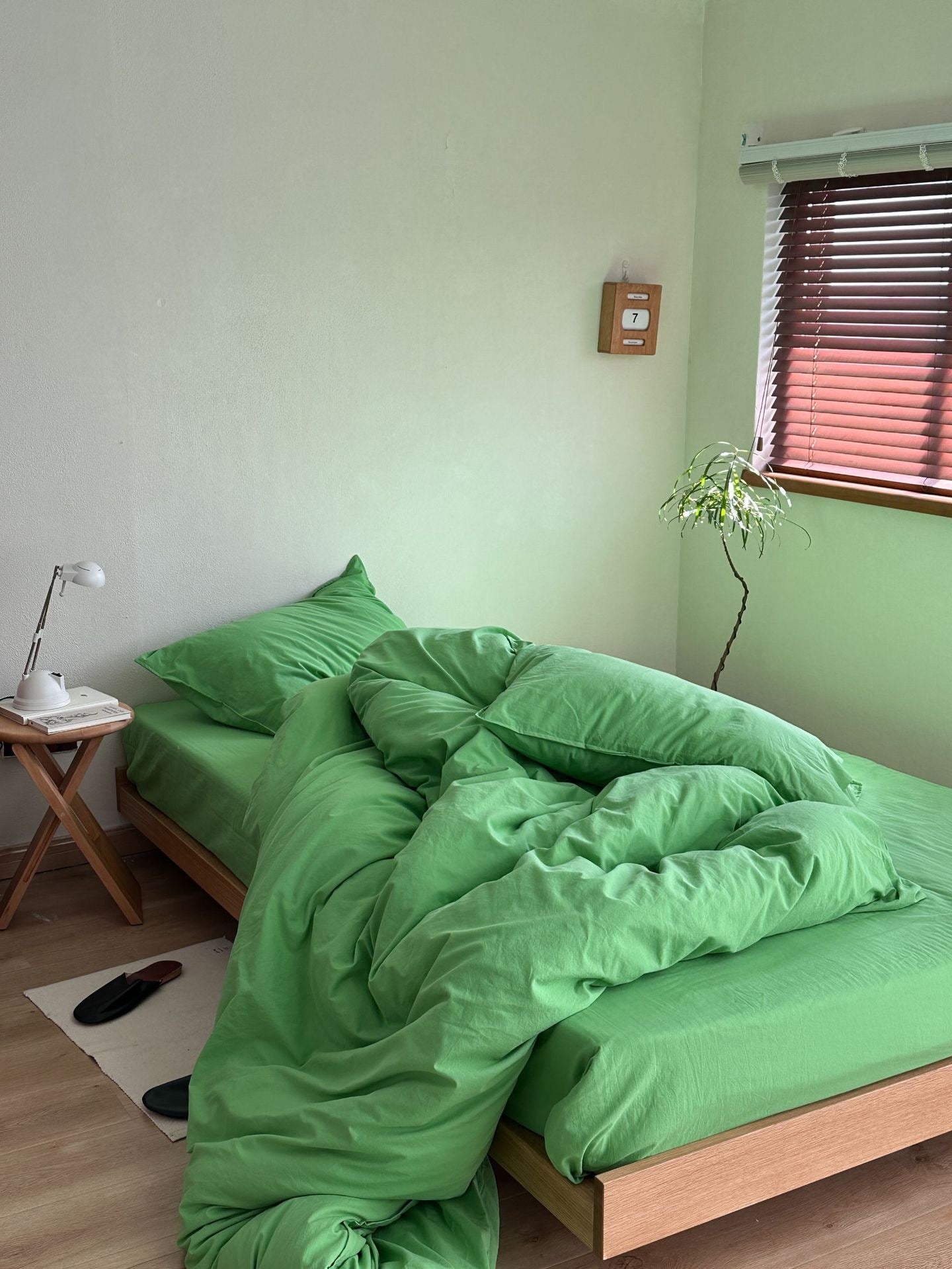A bed dressed in bright green bedding, including a duvet cover and matching pillowcases, styled in a minimalist bedroom with light green walls, a wooden side table with a lamp, and a window with brown blinds.