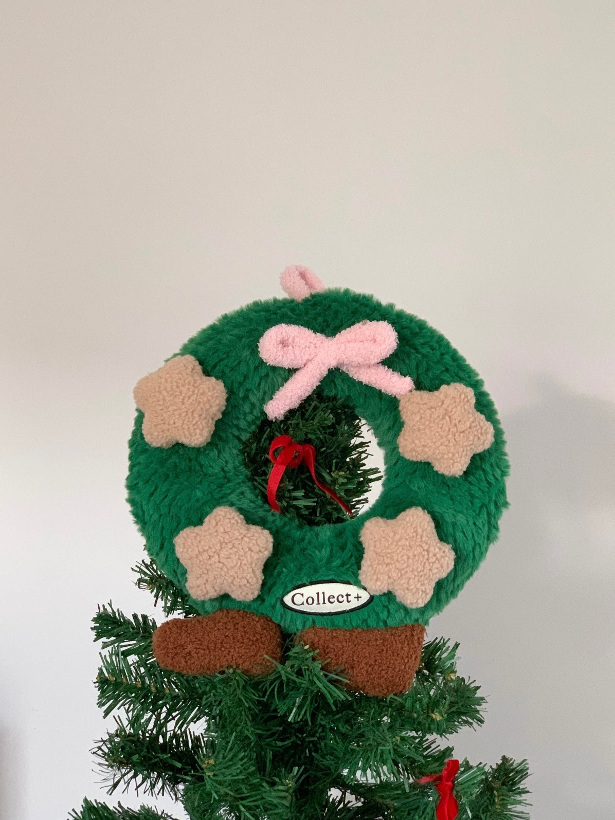 Green Christmas wreath cushion with pink bow displayed beside a Christmas tree