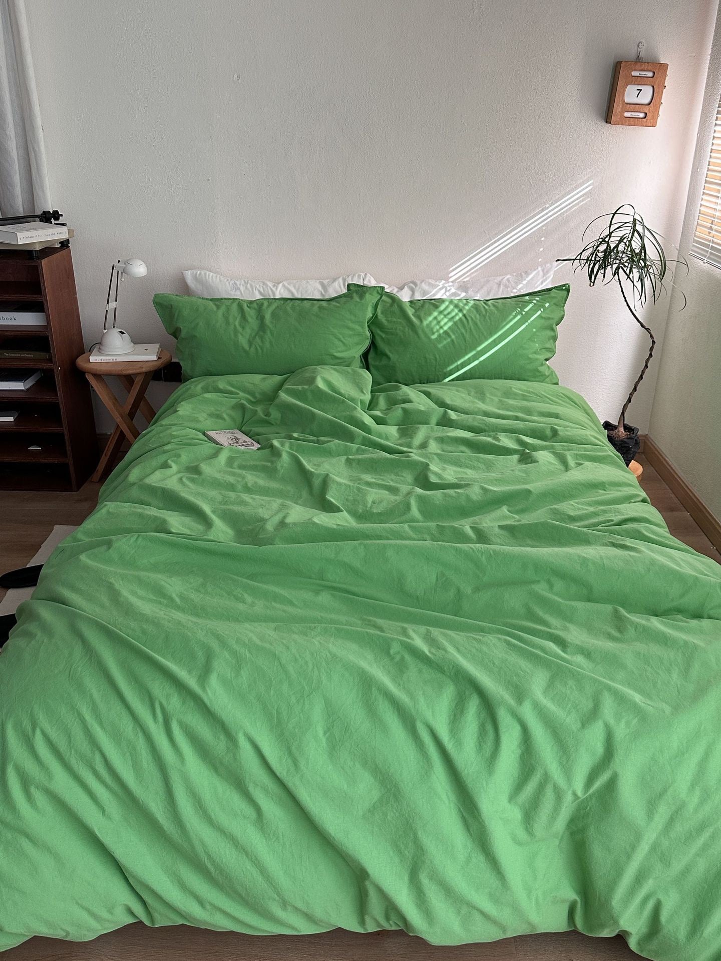 A bed made up with a bright green duvet cover and matching green pillows in a real bedroom setting, with a wooden nightstand, lamp, plant, and wall decor visible.