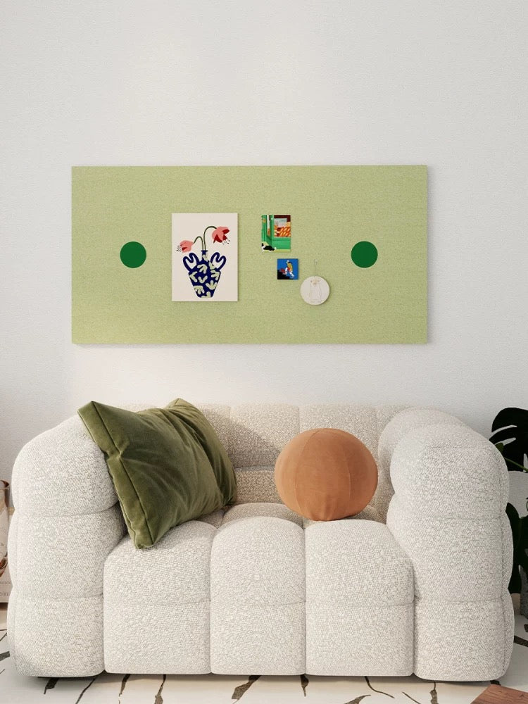 Green felt bulletin board displaying postcards above a white sofa with an orange cushion.