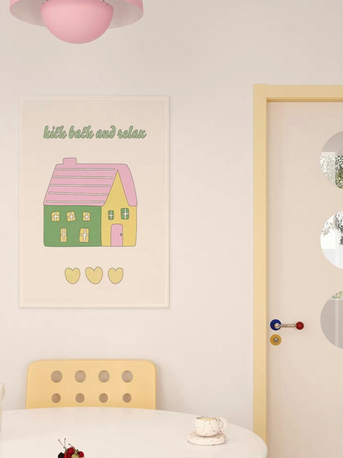 A green house wall hanging with a pink roof and yellow hearts is shown above a yellow dining chair.