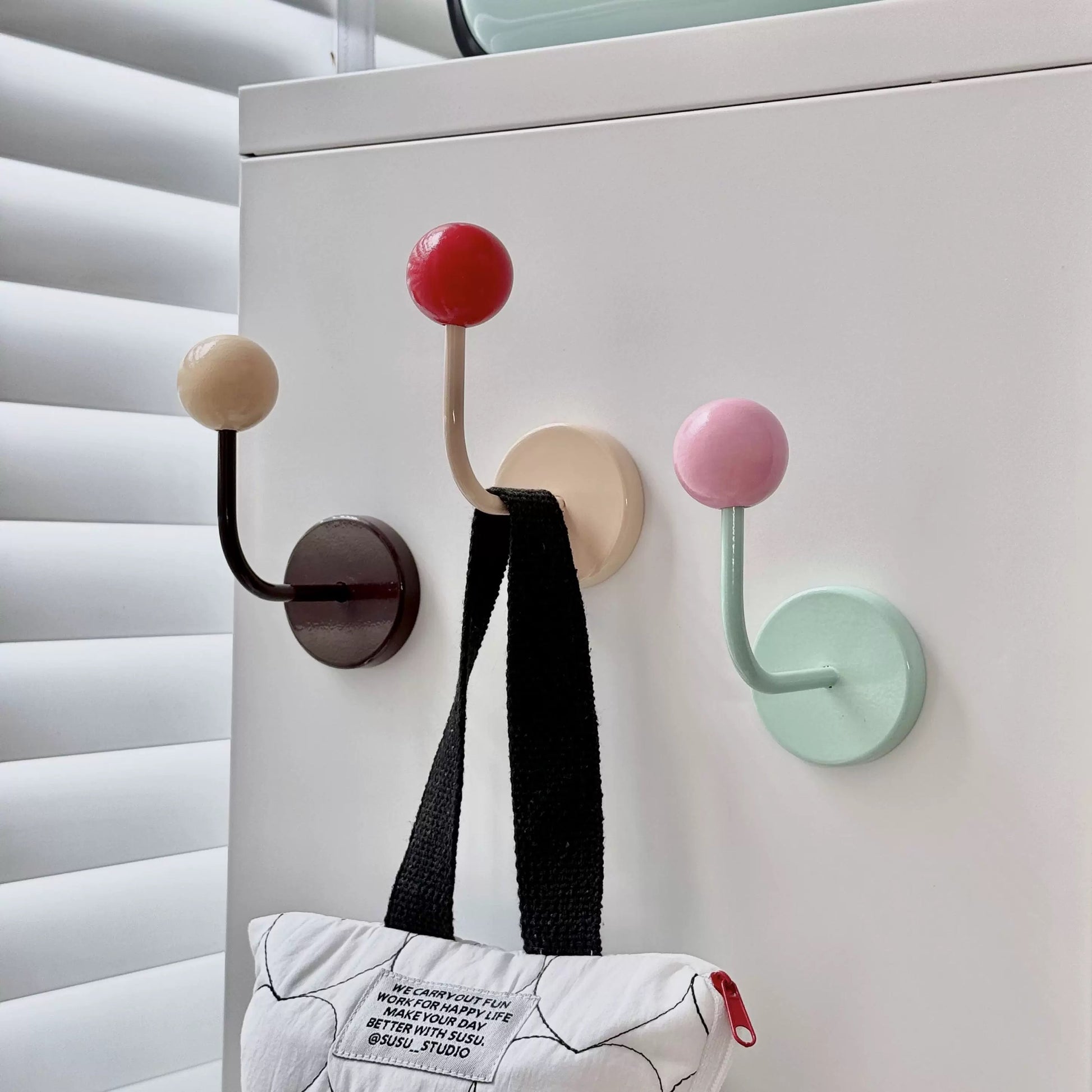 Three pastel-colored magnetic wall hooks with spherical handles hang on a white refrigerator door. The hooks are in dark brown, beige, and mint green, with red, cream, and pink spherical tips.