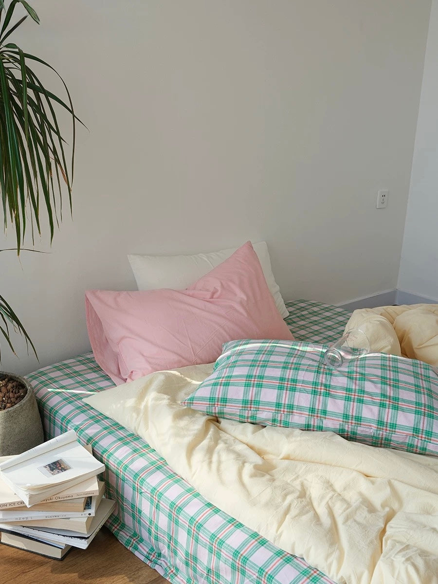 Bed with green and white checkered duvet, pink pillow, and books on a wooden floor.