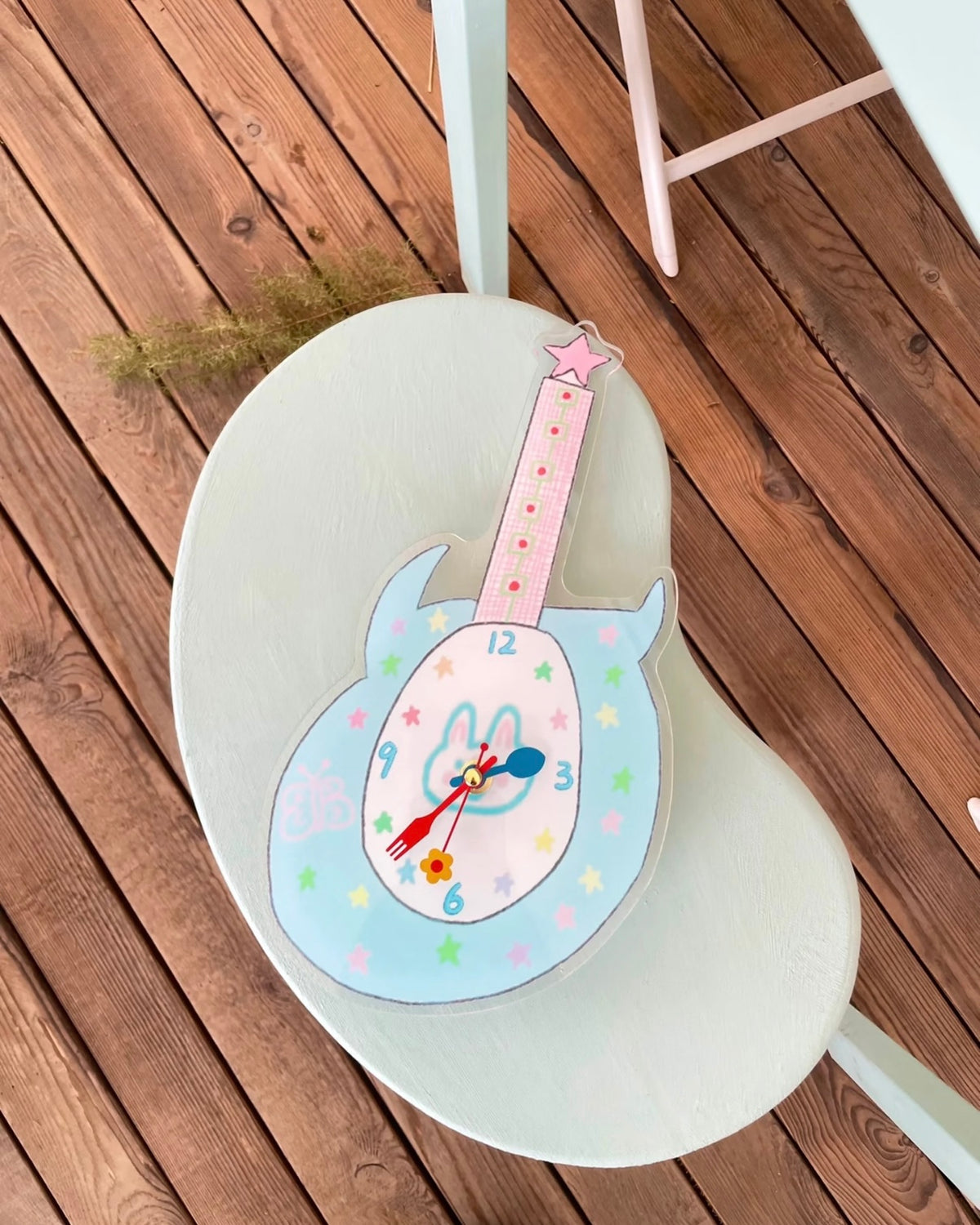 A decorative guitar-shaped wall clock with a bunny face and pastel star details, displayed on a white chair over a wooden deck floor.