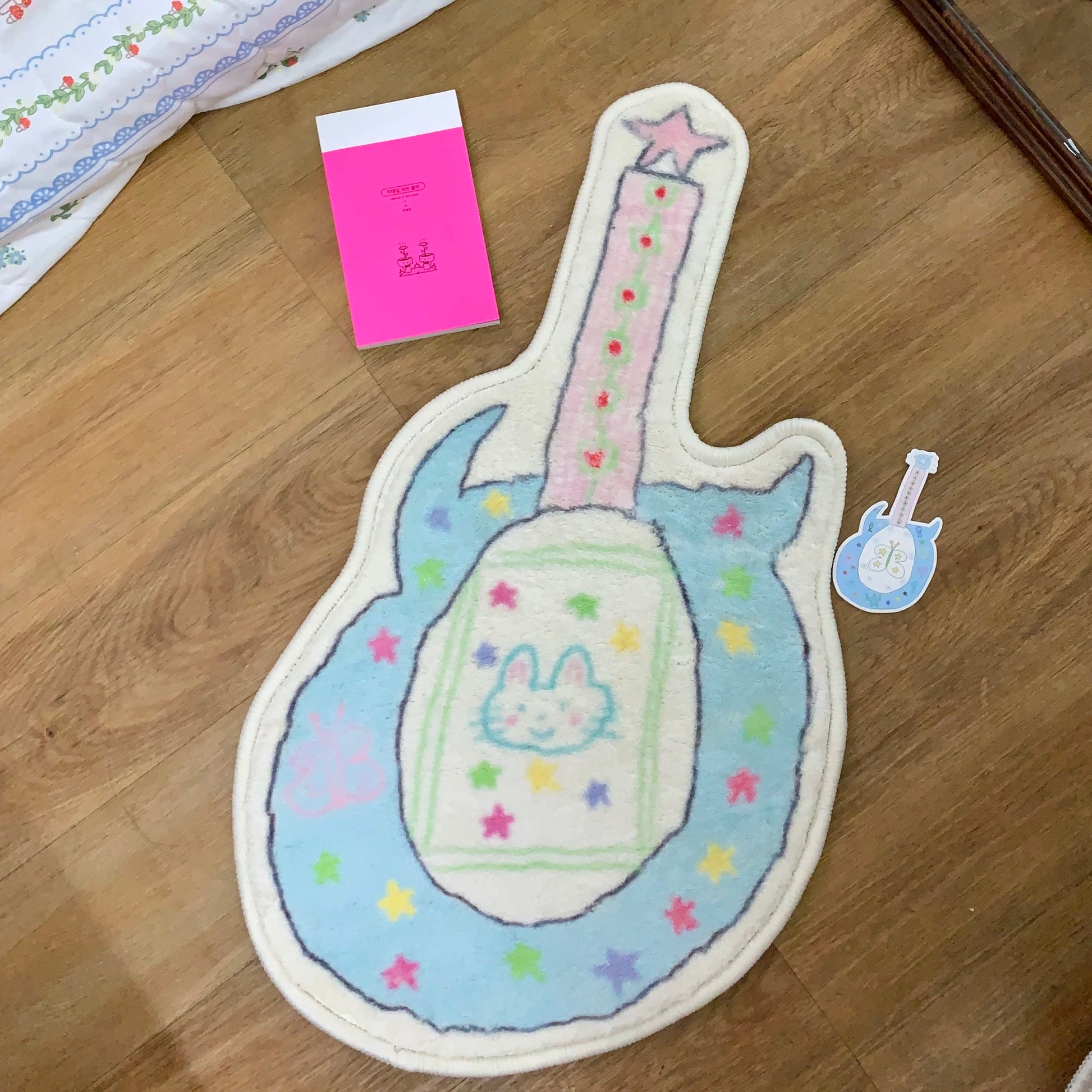 A guitar-shaped plush rug or mat in pastel blue, pink, and cream, decorated with colorful stars and a bunny face in the center, shown on a wooden floor next to a small matching sticker and a pink card.