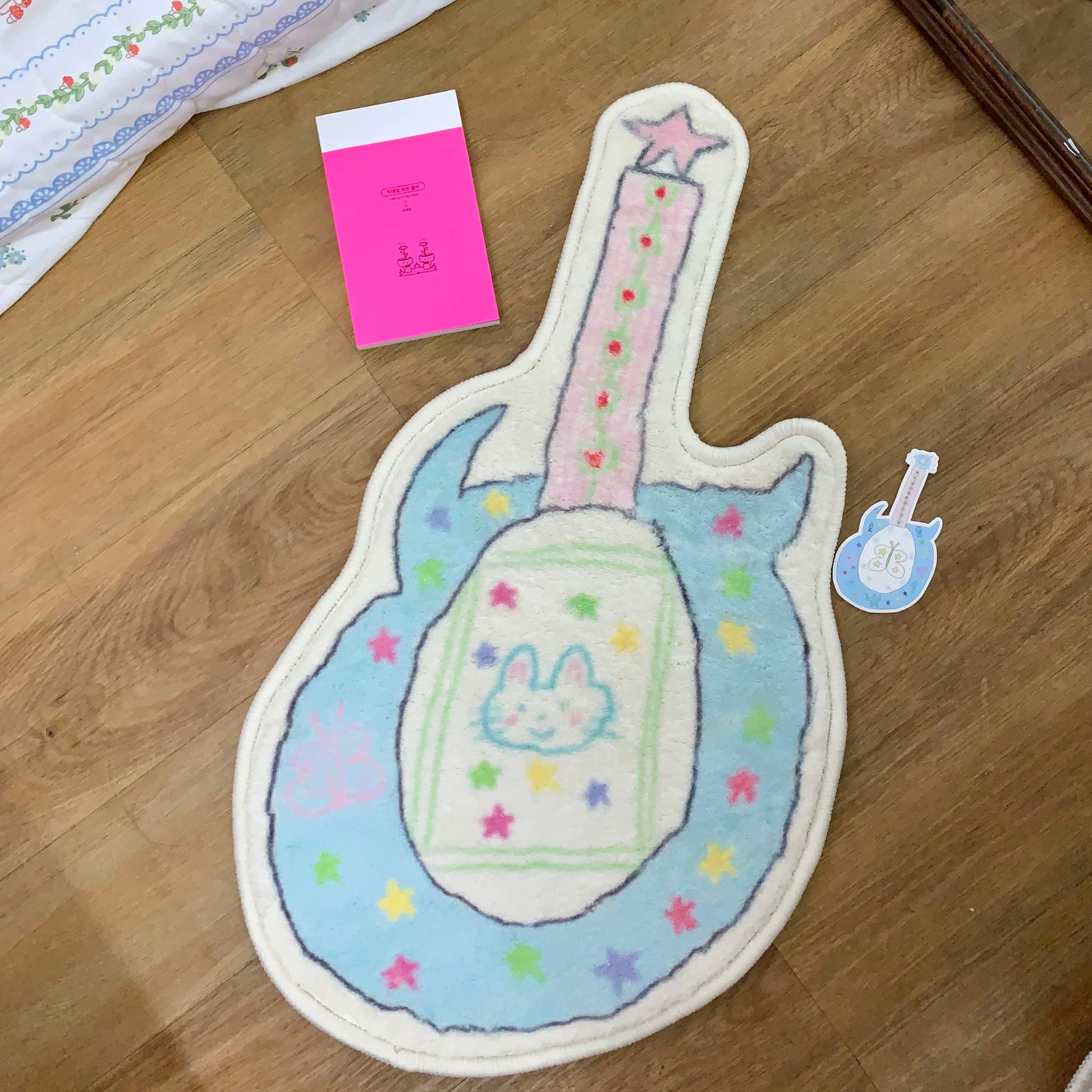 A guitar-shaped plush rug or mat in pastel blue, pink, and cream, decorated with colorful stars and a bunny face in the center, shown on a wooden floor next to a small matching sticker and a pink card.