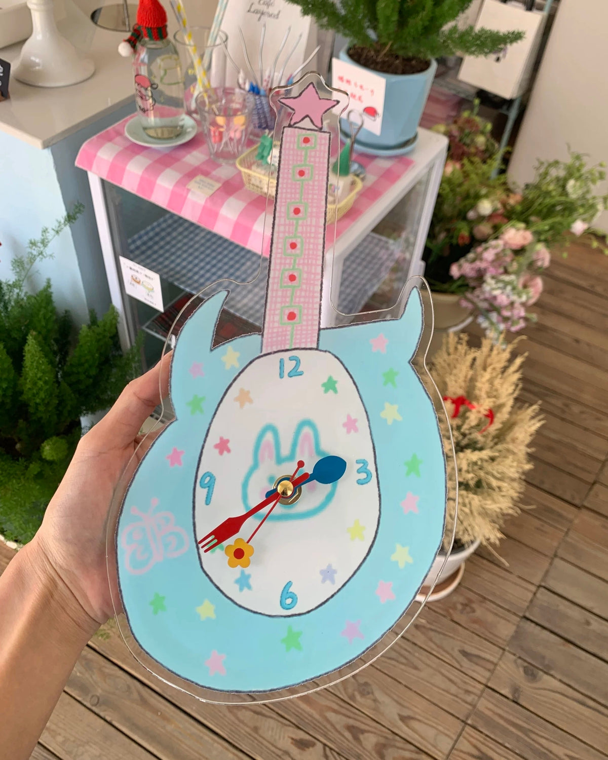 A hand holds a guitar-shaped wall clock with a light blue frame, pink neck, and a white center featuring a blue bunny outline and multicolored star accents. The background shows a decorated indoor space with a pink-and-white checkered table, plants, and wooden flooring.