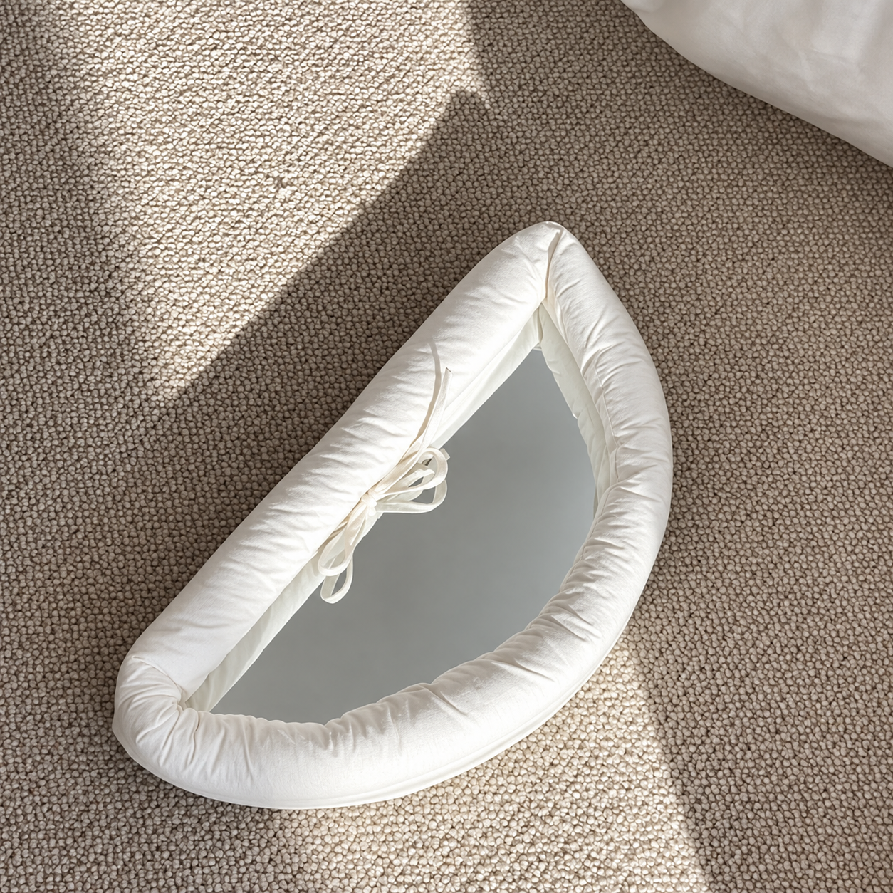 A crescent-shaped white padded cushion with a mirror inset, styled on a textured beige carpet with soft natural shadows.