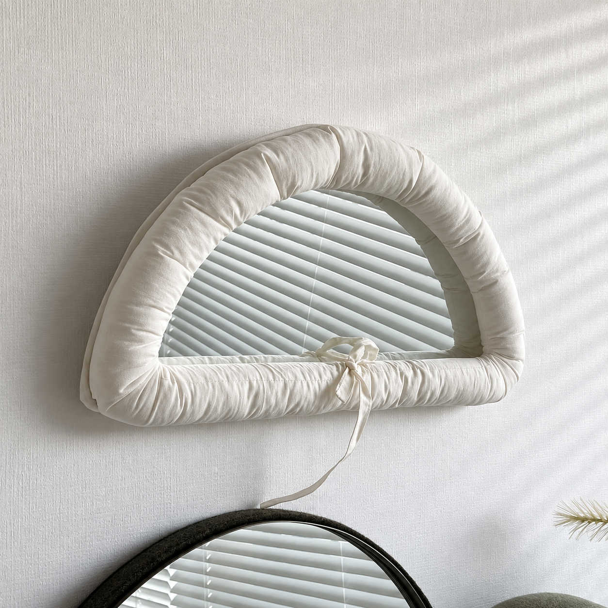 A padded cream-colored arch-shaped mirror with a tie detail, mounted on a textured white wall with soft diagonal shadows.