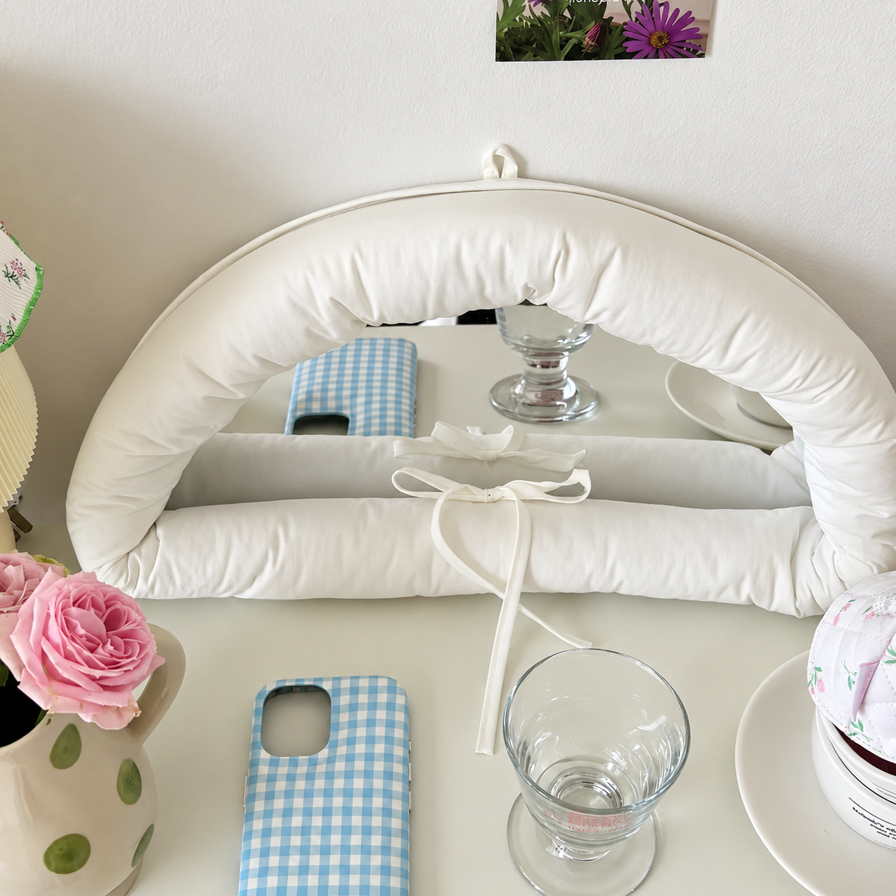 A white padded arch-shaped cushion or pillow with tie straps, displayed on a tabletop with glassware, a pink rose in a vase, and a blue-and-white gingham phone case nearby.