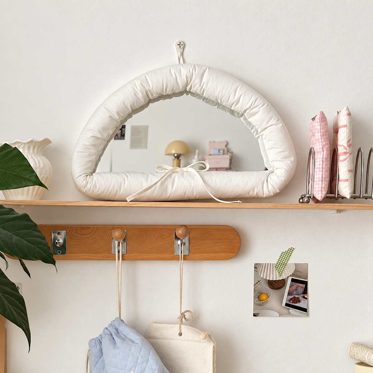 A padded arched wall mirror with a cream fabric frame and bow detail, mounted on a light wood shelf in a styled interior setting.