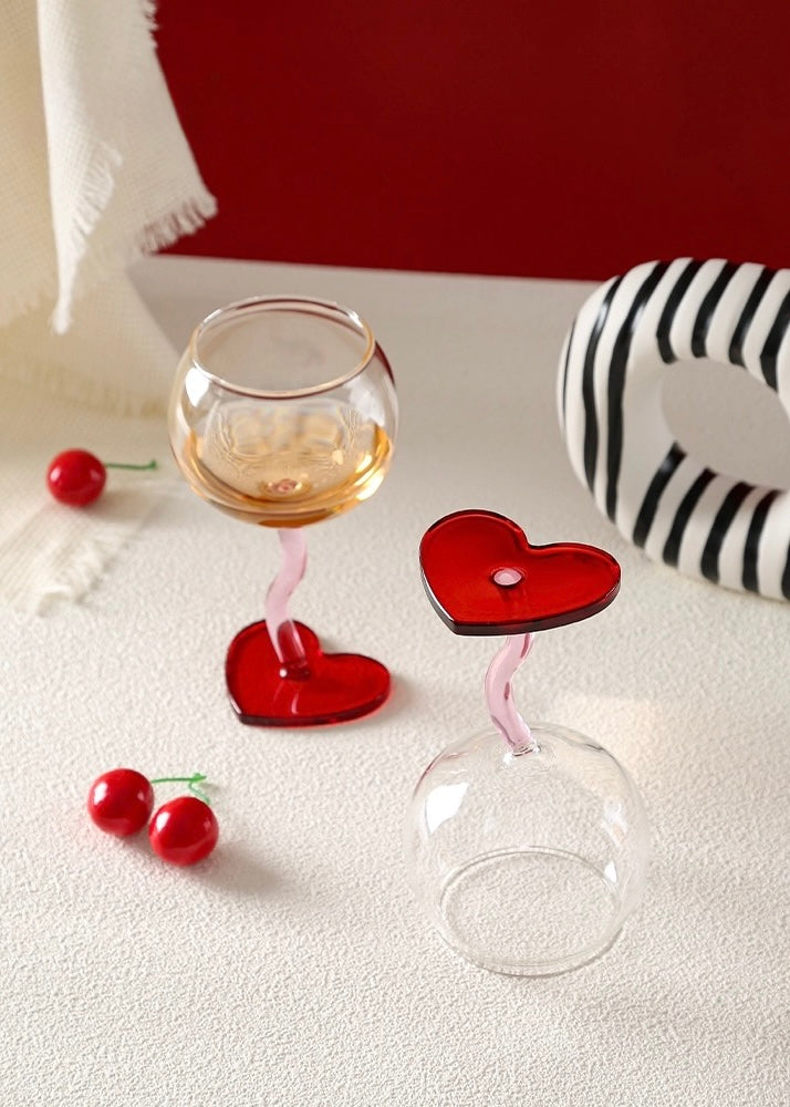 Two heart-shaped wine glasses with red cherries on a white surface.