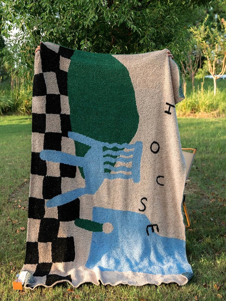 A large woven throw blanket with a bold abstract pattern is draped outdoors on grass in front of trees. The blanket features a black-and-beige checkerboard section, a large dark green shape, a light blue chair motif, and scattered black letters on a beige background.