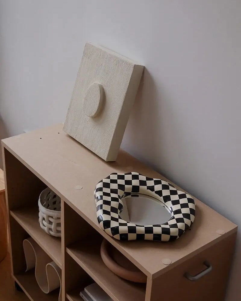A beige rectangular wall-mounted wooden box with a circular cutout, leaning against a white wall on top of a light wood storage cabinet. A black-and-white checkered oval cushion rests on the cabinet, and a white woven basket is visible in an open shelf compartment.