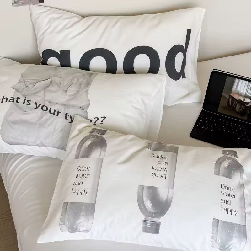 White bedding set with printed graphic pillows on a bed, featuring black text and a grayscale water bottle design, alongside a black laptop on a bedside table.
