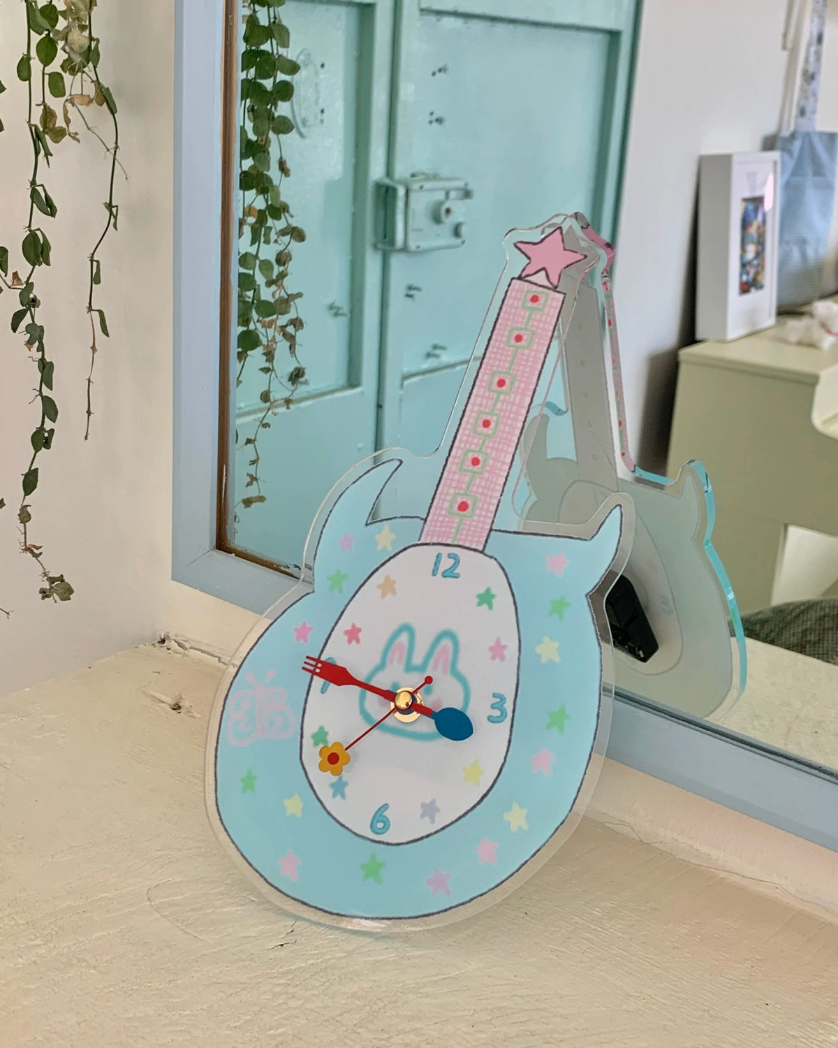 A guitar-shaped wall clock with a pastel blue body, pink neck, and a white rabbit face on the face, styled on a windowsill with a mirror and green trailing plants in the background.