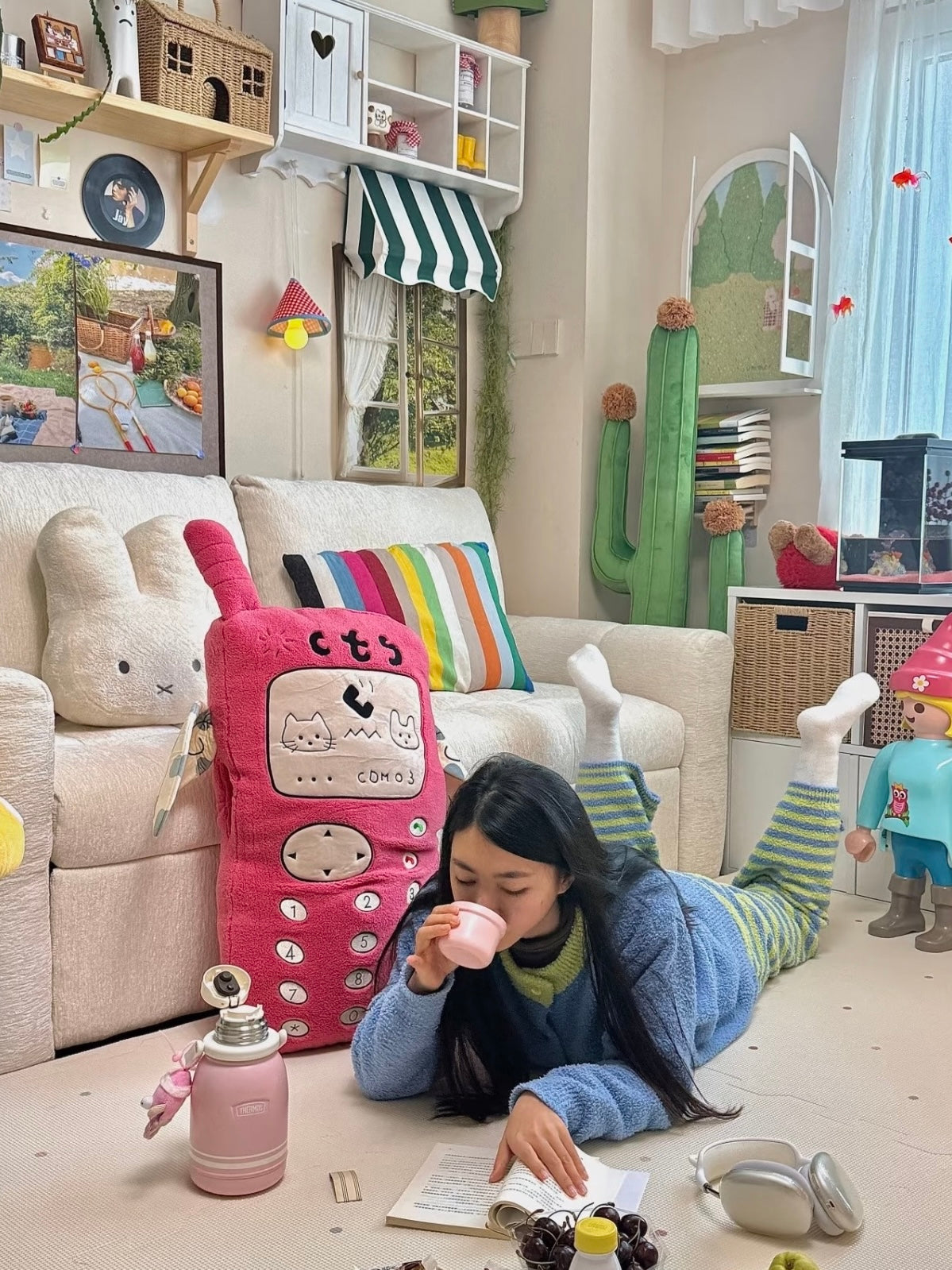 A pink Kawaii phone pillow stands near a sofa while a person lies on the floor beside it, with the keypad facing out.