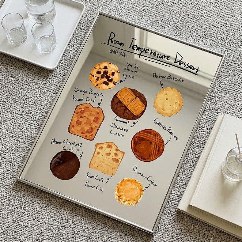 A rectangular mirrored tray with a printed dessert menu titled 'Room Temperature Dessert' lies on a textured gray fabric surface. The tray reflects cookies, cakes, and pastries, and is styled with small glass cups and a book nearby.
