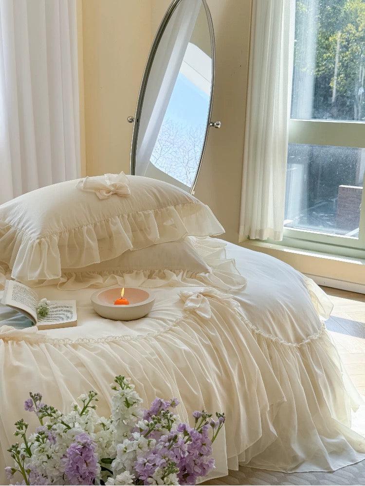 Delicate white bedding with ruffled edges on a bed, featuring a candle and flowers in a softly lit room.