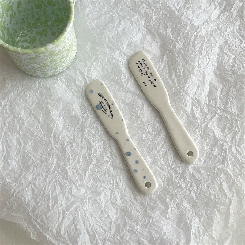Two white ceramic spatulas with blue speckles and black printed text, displayed on a crumpled white paper background next to a matching green and white speckled ceramic cup.