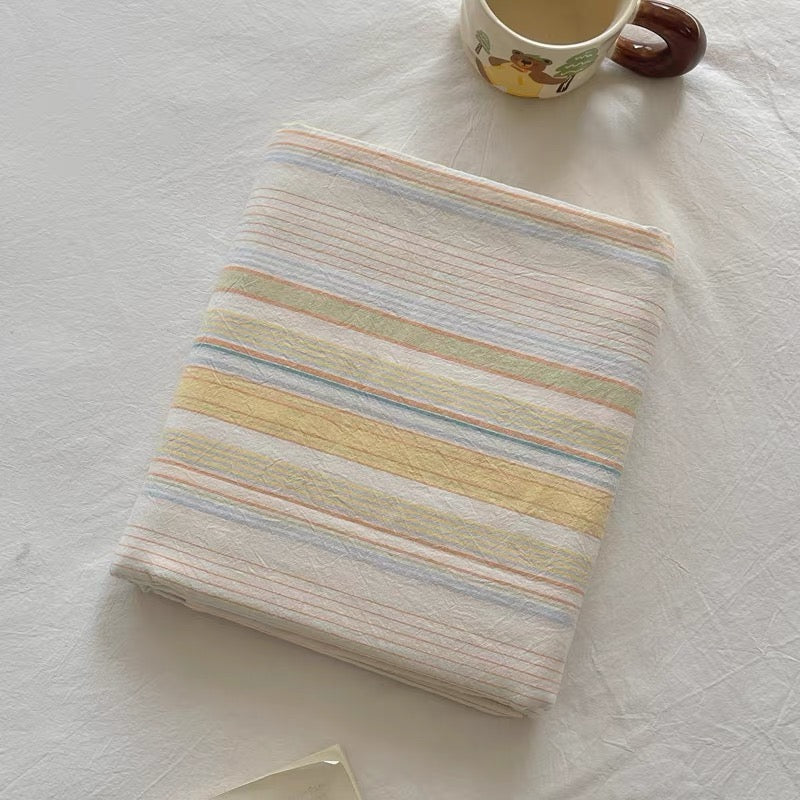 A folded striped fabric item, likely a blanket or throw, with pastel horizontal stripes in cream, yellow, light blue, and pale green, styled on a white textured fabric surface next to a ceramic mug.