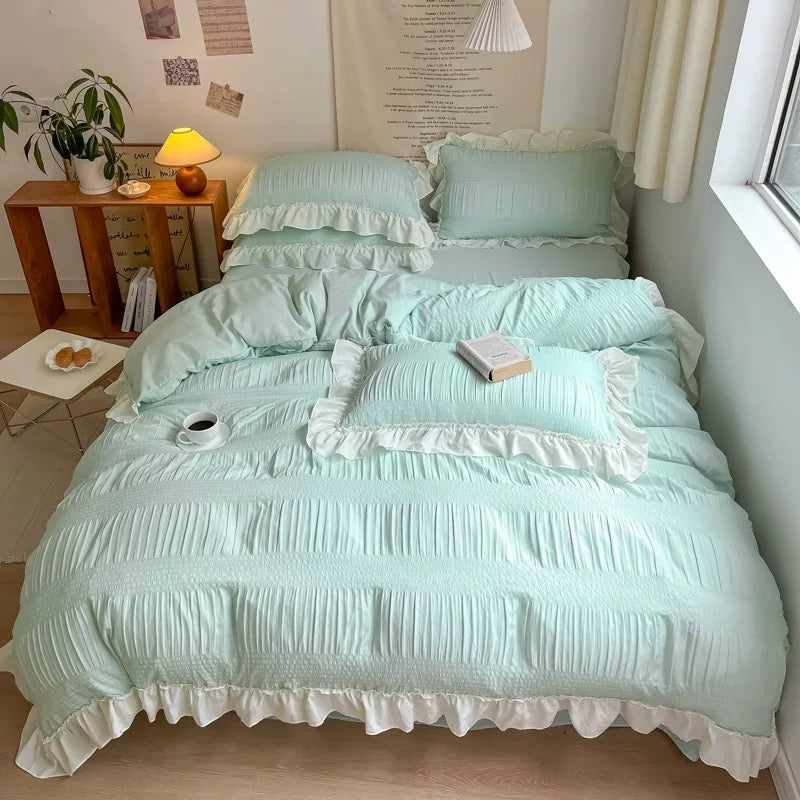 Light blue bedding set with ruffled edges on a bed in a room with a lamp and plant.