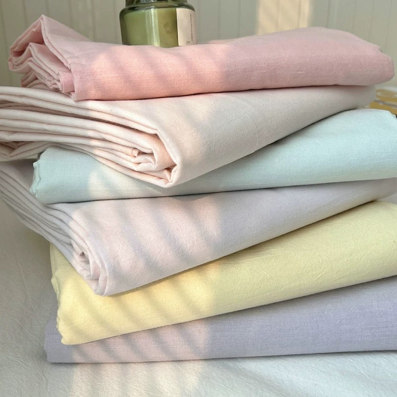 A stack of folded pastel fabric sheets or linens in pink, cream, pale blue, lavender, and pale yellow, photographed indoors with soft natural light and a blurred green bottle in the background.