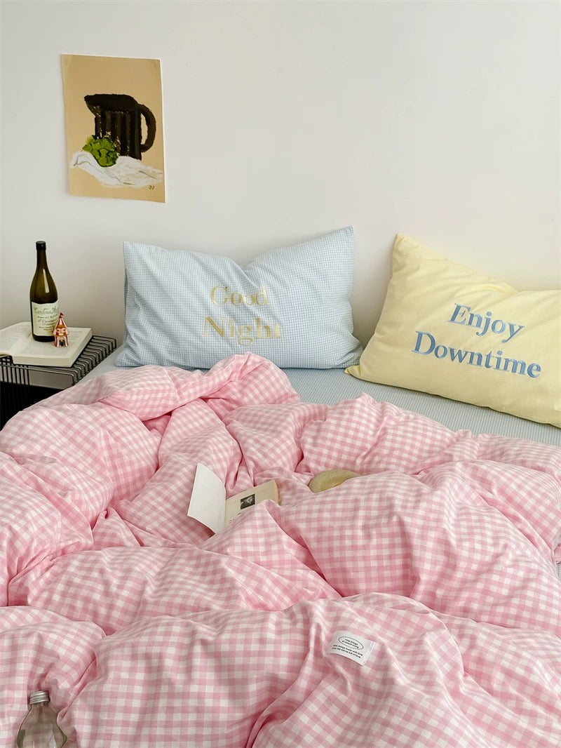 Pink checkered comforter on a bed with pillows and a bottle of wine on a nightstand.