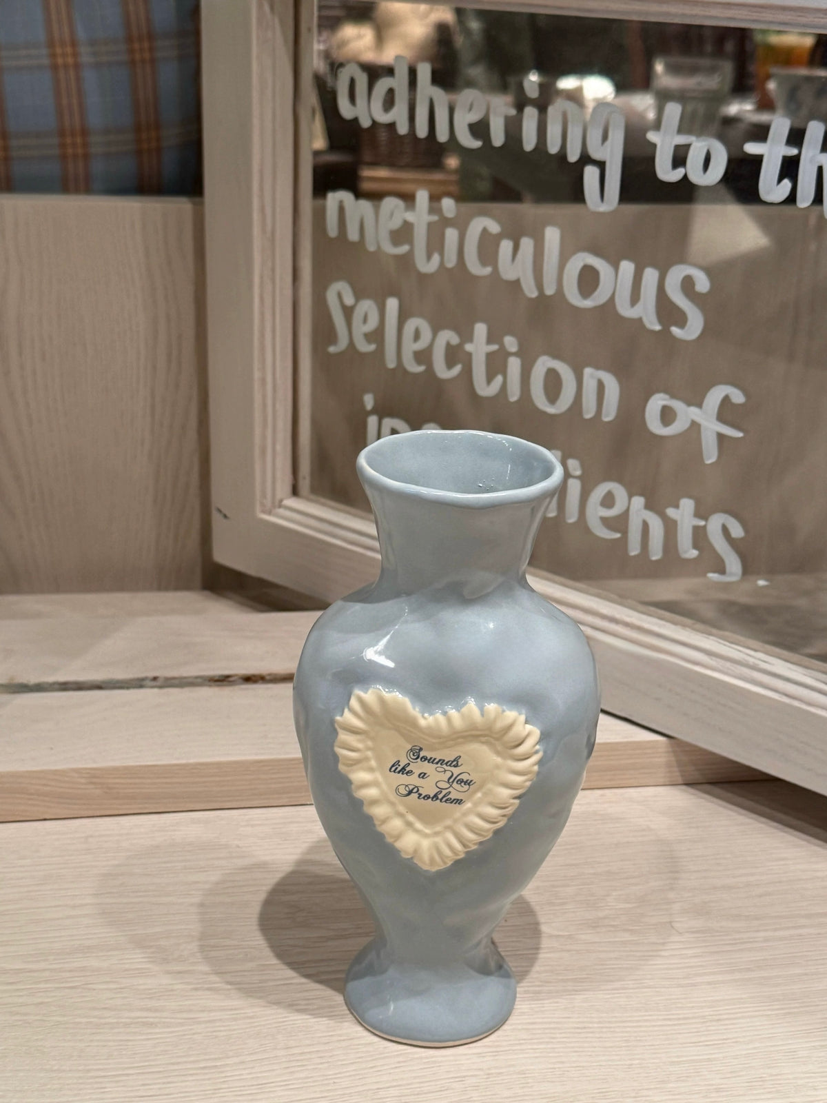 Light blue ceramic vase on a wooden tabletop, with a raised white heart, blue lettering and a flared neck.
